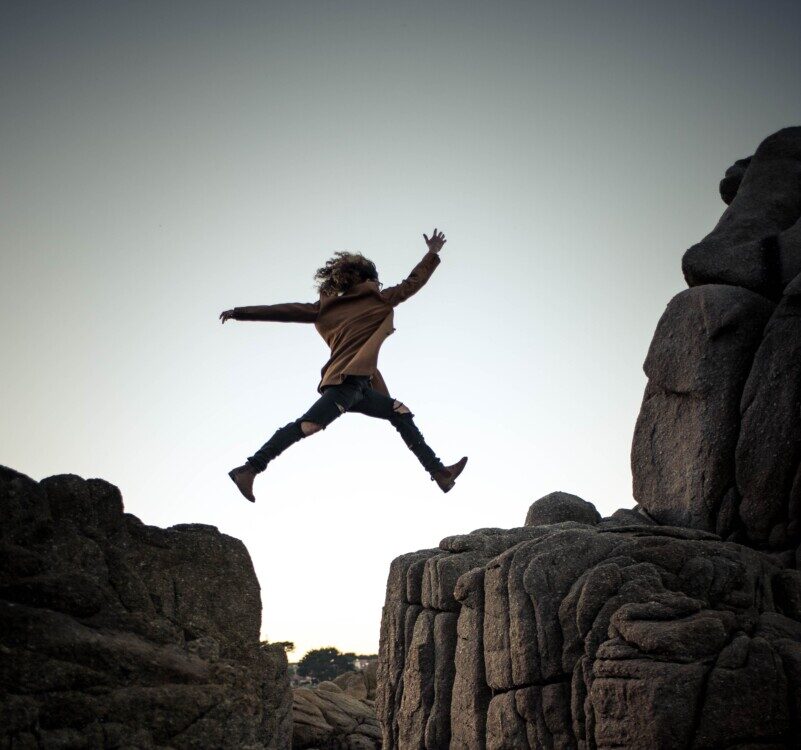 person jumping on big rock under gray and white sky during daytime